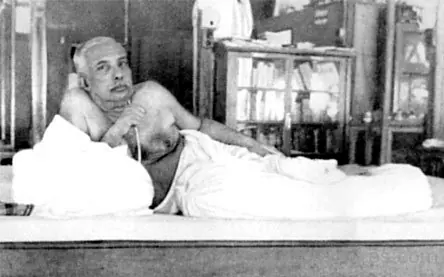 Sri Sri Thakur Anukulchandra in his later years, reclining on his bed against a pillow, bookshelves behind