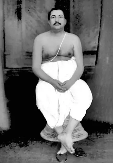 Young Sri Sri Thakur Anukulchandra seated on a cane stool in a white dhoti, hands folded in his lap