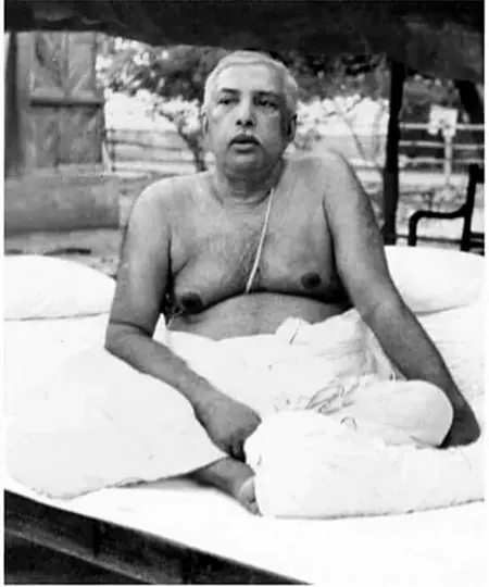 Sri Sri Thakur Anukulchandra in his later years, seated on a bed set out in the open beneath a canopy