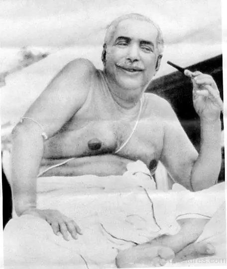 Sri Sri Thakur Anukulchandra in his later years, smiling on his bed with a pipe in his raised hand