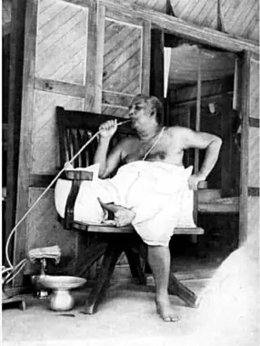Sri Sri Thakur Anukulchandra seated in a wooden chair on a verandah, smoking his hookah through a long pipe